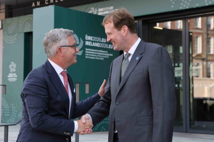 Two men shake hands and laugh at the entrance of the Irish presidency’s meeting venue at Dublin Castle, in front of green ÉIRE2026.EU branding.