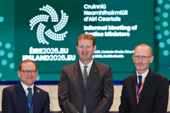 Three men stand in front of a large green screen reading “Informal Meeting of Justice Ministers” with the Irish presidency’s ÉIRE2026.EU logo.