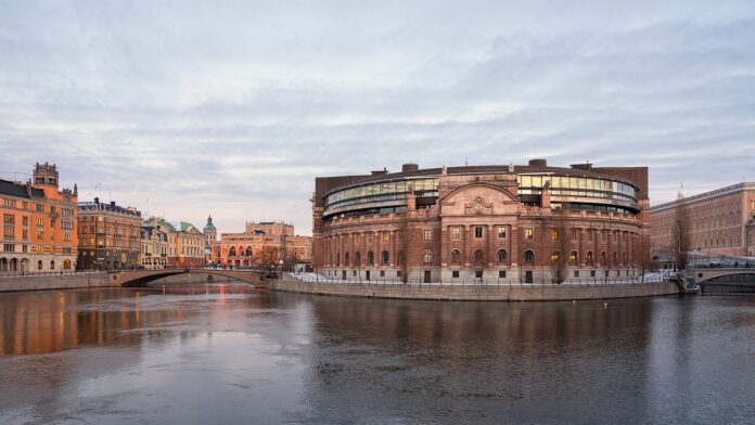 The Swedish Parliament House in Stockholm, a curved stone building with a glass top storey, seen across the water on a winter afternoon